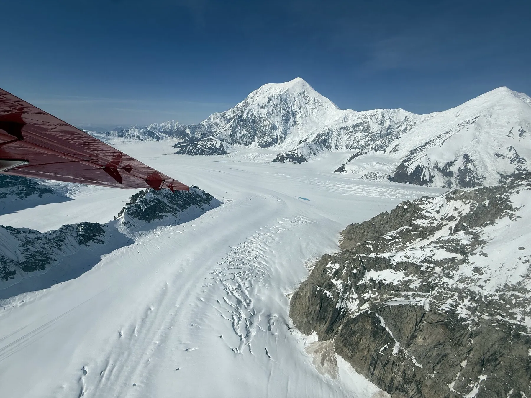 A glacier winding between snowy mountains, photographed beneath the wing of a small plane